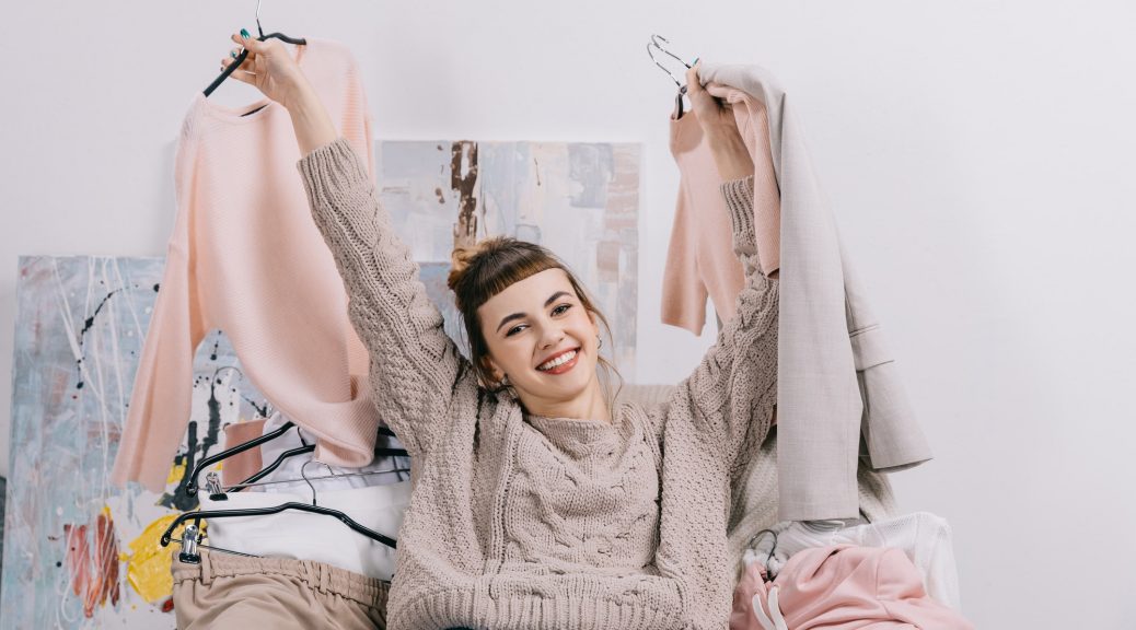 smiling girl sitting on armchair and holding hangers with clothes in hands